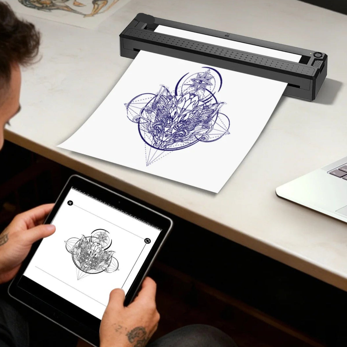 Black thermal tattoo stencil printer on a worktable beside a printed design and a tablet used to prepare artwork